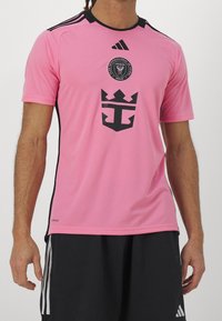 Mies, joka pukeutuu vaaleanpunaiseen Adidas-jalkapallopaitaan, jossa on Club Internacional de Futbol Miami -logo, ja mustiin shortseihin, joissa on valkoiset raidat.