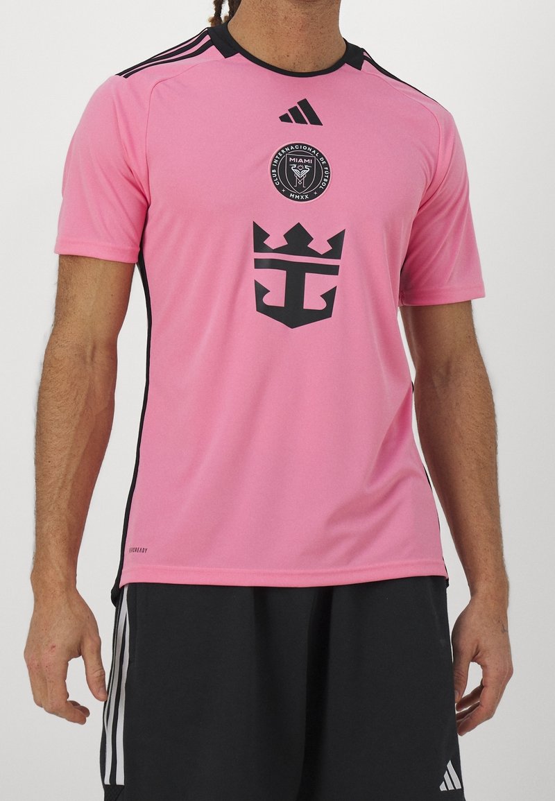 Mies, joka pukeutuu vaaleanpunaiseen Adidas-jalkapallopaitaan, jossa on Club Internacional de Futbol Miami -logo, ja mustiin shortseihin, joissa on valkoiset raidat.