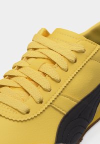 Κίτρινο αθλητικό παπούτσι με υφασμένα κορδόνια και μαύρο σουέντ λεπτομέρειες κοντά στην σόλα, εμφανίζεται σε κοντινή λήψη.