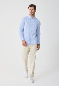 Light blue long-sleeve button-up shirt in linen fabric, paired with cream trousers and white sneakers. Simple collar and fitted design.