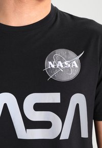 Camiseta de algodón negra con un logotipo redondo de la NASA en gris y blanco con un diseño de cohete. Gran texto "NASA" en plata debajo del logotipo.