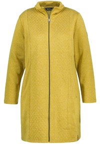 Abrigo amarillo de cremallera con cuello mandarín, mangas largas, tejido texturizado, bolsillos verticales y acentos en patrón de diamante en los laterales.