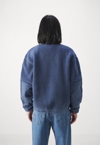 Marinblå fleece-tröja med texturerad tyg, oversized silhuett och kontrasterande blå ärmar. Parat med ljusa denimjeans.
