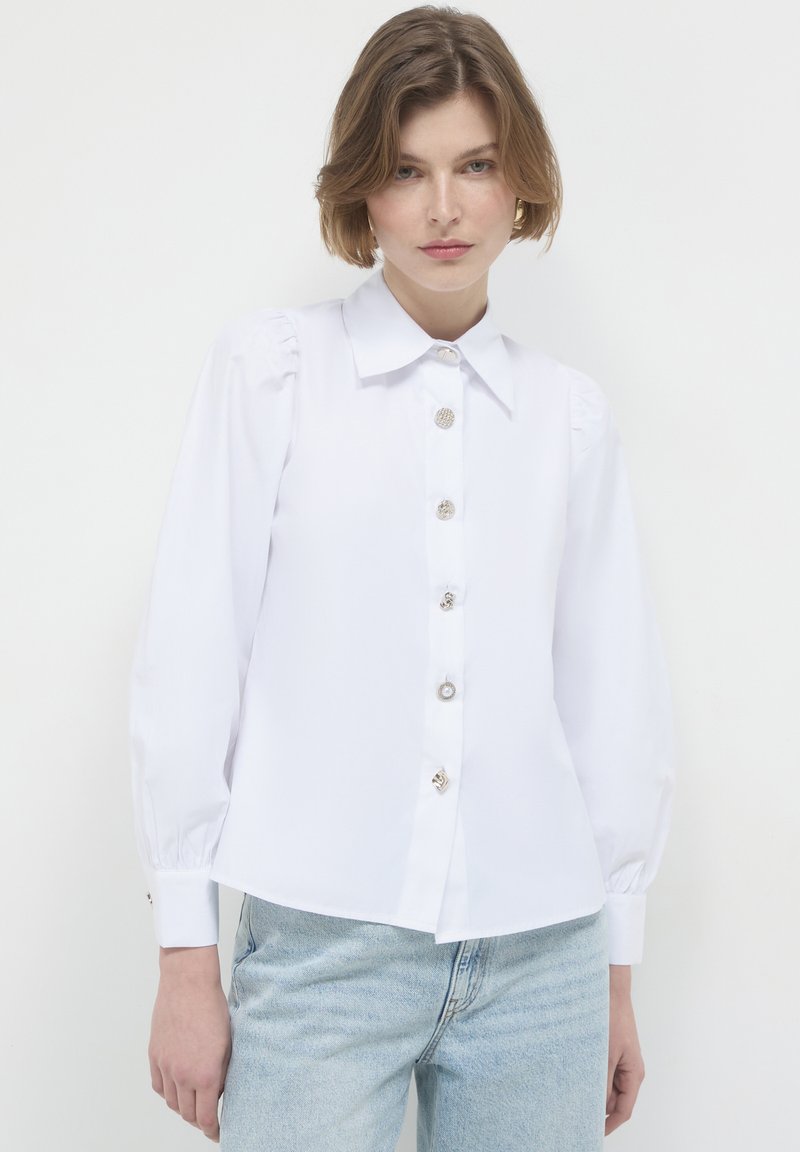 Woman wearing a white button-up blouse with large decorative buttons and light blue jeans against a plain white background.