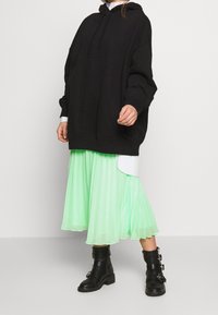 Felpa nera oversize, gonna plissettata verde menta, stivaletti neri alla caviglia con fibbie. Maglietta bianca visibile all'orlo. Stile di sovrapposizione informale.