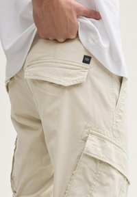 Beige Cargo-Shorts mit mehreren Taschenakzenten, einer glatten Textur und einem kleinen schwarzen Etikett auf der Rückseite, die eine lässige Passform bieten.