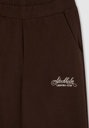 Pantaloni della tuta marroni con una cintura elastica, dotati di tasche laterali e un logo ricamato bianco con la scritta "Stockholm Leisure Club."