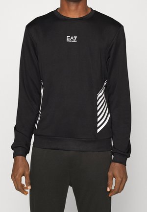 Mann trägt einen schwarzen EA7 Emporio Armani Sweatshirt mit langen Ärmeln und weißen schrägen Streifen an den Seiten sowie schwarze Hose.