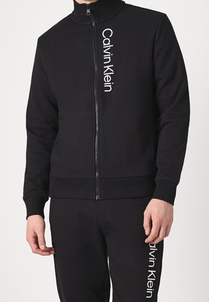 Muž v černé bundě na zip a odpovídajících kalhotách, obojí s vertikálními bílými logy "Calvin Klein" na levé straně.