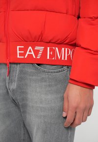 Tuvumā redzams cilvēks, kurš valkā sarkanu pūkainu jaku ar EA7 Emporio Armani logotipu uz jostas daļas un gaiši pelēkus džinsus.