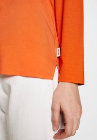 Nahaufnahme einer Hand, die an weißen Hosen ruht, getragen mit einem orangen Langarmshirt, an dessen Saum ein kleines Stoffetikett angebracht ist.