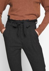 Black tapered pants with a textured fabric, featuring a tied waistband and side pockets, paired with a brown knit sweater.