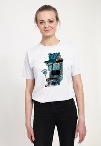Bílý bavlněný tričko s barevným grafickým designem budovy s modrými a červenými akcenty. Model je nosí s černými džíny.
