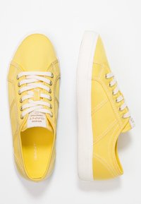 Gula kanvas sneakers med vita gummisulor, snören och sömmar. Har en logotapp på sidan och en texturerad ovandel.