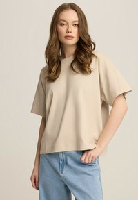 Beżowy, oversize'owy t-shirt wykonany z bawełny, z krótkim rękawem i okrągłym dekoltem, zestawiony z jasnoniebieskimi dżinsami. Minimalistyczny design.