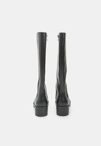 Un paio di stivali alti in pelle nera con tacco a blocco visti da dietro su uno sfondo chiaro e uniforme.