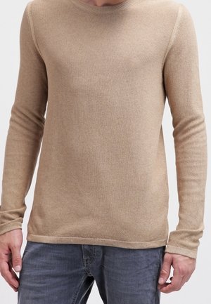Beige gestrickter Pullover mit langen Ärmeln und rundem Hals, der ein strukturiertes Muster und gerippte Bündchen aufweist. Getragen mit blauen Jeans.
