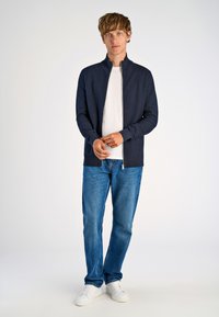 Marinblå zip-up jacka i texturerat tyg, matchad med en vit t-shirt, blå jeans och vita sneakers. Enkel bakgrund.