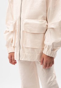 Světle růžová bunda na zip se dvěma předními kapsami, žebrovanými manžetami a jemnou vzorovanou texturou. Doplněna odpovídajícími kalhotami.