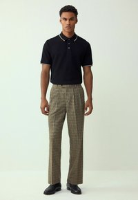 Schwarzes Polohemd mit kontrastierendem Kragen, kombiniert mit braunen und beigen karierten Hosen. Das Modell steht in schwarzen Schuhen vor einem hellen Hintergrund.