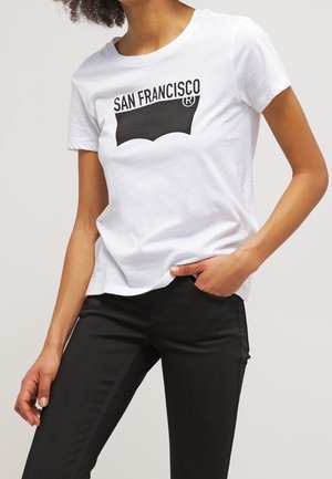 Persona con camiseta blanca con el texto "SAN FRANCISCO" sobre un rectángulo negro, combinada con pantalones negros.