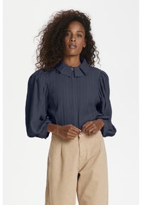Navyblaue strukturierte Bluse mit Kragen und Puffärmeln, die über eine Knopfleiste an der Vorderseite verfügt. Getragen mit cremefarbenen, hoch taillierten Hosen.