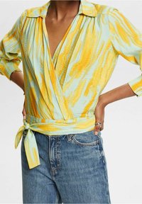 Haut portefeuille à col en bleu clair avec des rayures abstraites jaunes et oranges. Manches longues, ceinture à nouer à la taille, tissu texturé. Porté avec un jean bleu.