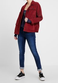 Červená kordurová bunda s knoflíkovým zapínáním a předními kapsami, kombinovaná s tmavě modrými skinny džíny a černými teniskami.