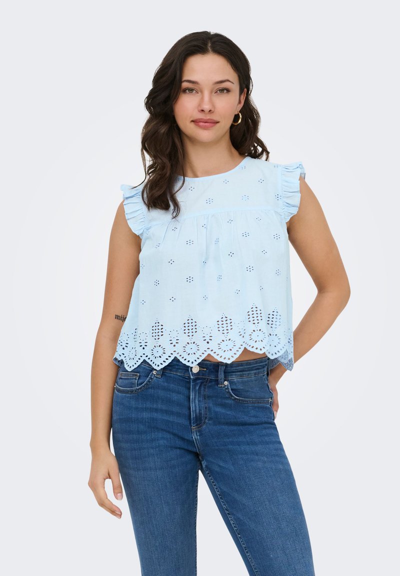 Femme aux cheveux bruns ondulés portant un haut bleu clair sans manches à œillets avec des volants et un jean bleu, debout avec une main sur la hanche.