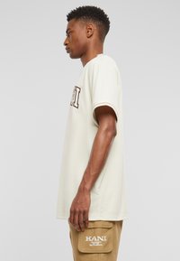 Lys beige kortærmet t-shirt med rund hals og mørkebrun logo på brystet, vist på en model fra siden.
