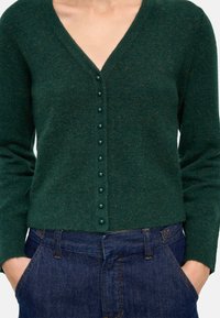 Zielony kardigan z wełny w mieszance z dekoltem w kształcie litery V, ozdobiony rzędem ciemnozielonych guzików z przodu. W zestawie z ciemnoniebieskimi dżinsami.