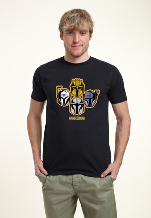 Ung mand iført en sort t-shirt med farverige Mandalorian-hjelme og khakibukser, stående mod en lys baggrund.