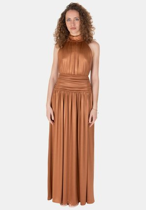 Donna dai capelli ricci che indossa un abito lungo in raso color rame con scollo halter, in piedi davanti a uno sfondo chiaro e semplice.