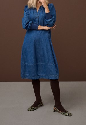 Vrouw met een blauwe denim jurk tot op de knie, zwarte panty's en slingback schoenen met lage hak en slangenhuidpatroon, staand met gekruiste armen.