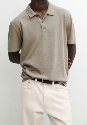 Homme portant un polo beige à manches courtes rentré dans un pantalon blanc cassé avec une ceinture tressée foncée, debout devant un fond uni.