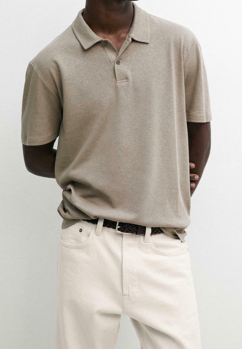 Homme portant un polo beige à manches courtes rentré dans un pantalon blanc cassé avec une ceinture tressée foncée, debout devant un fond uni.