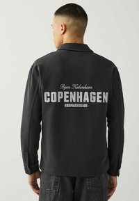 Schwarze Jacke mit einem Kragen-Design. Auf der Rückseite ist gedruckter Text: "Byen København COPENHAGEN KØBMAGERGADE" zu sehen, mit geknöpften Manschetten.
