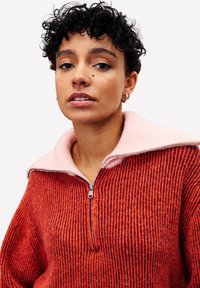 Un pull zippé chic avec une texture côtelée en orange et rouge, contrasté par un col rose doux. Quincaillerie de zip remarquable au niveau du col.