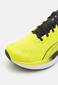 Zapatilla de correr amarilla brillante con parte superior de malla, cordones negros y suela Texturizada. Presenta un diseño elegante con una línea de acento blanca y gris.