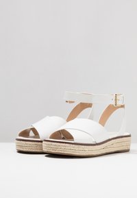 Valkoinen nahkainen espadrille-sandaali, jossa on ristikkäin muotoiltu yläosa, nilkkaremmi ja 3-tuumainen luonnollinen jutee-vuori. Suora pinta.