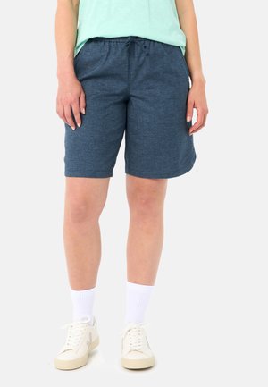 Person trägt marineblaue Shorts mit Kordelzug, hellmintgrünes Hemd, weiße Socken und weiße Turnschuhe vor einfachem Hintergrund.