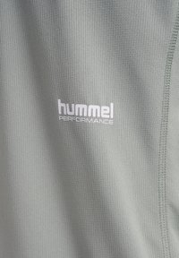 Tissu texturé gris clair avec gros plan sur le logo blanc "hummel PERFORMANCE" et couture visible sur le côté droit.