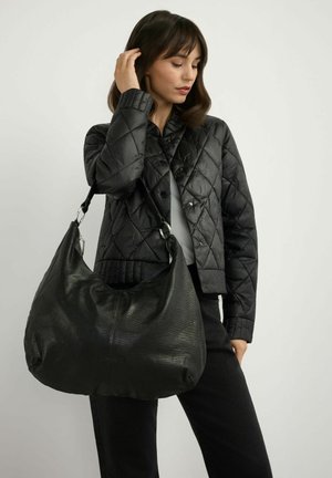 Kobieta w czarnej pikowanej kurtce i spodniach trzyma dużą czarną skórzaną torbę na ramię na szarym tle.