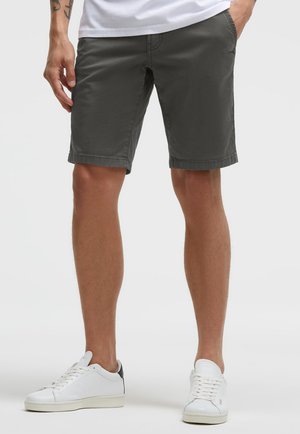 Mann trägt dunkelgraue knielange Shorts, weiße Turnschuhe mit schwarzer Ferse und weißes T-Shirt, eine Hand in der Tasche.