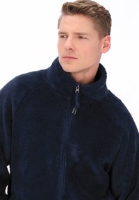 Tengerészkék polárdzseki álló gallérral és teljes cipzárral. Puha, texturált anyag raglán ujjal és finom varrási részletekkel.