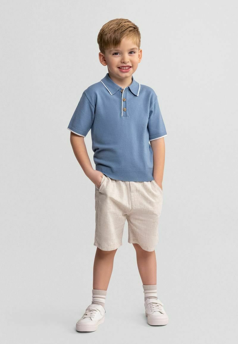 Młody chłopiec stojący z rękami w kieszeniach, ubrany w niebieską polo z krótkim rękawem, beżowe szorty, białe skarpetki i białe trampki, uśmiechający się do kamery.