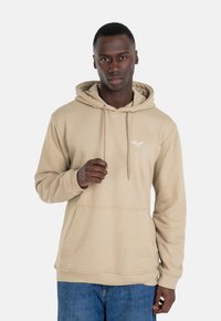 Beige Kapuzenpulli mit Kordeln, Fronttasche und gerippten Bündchen. Weicher Stoff mit minimaler Markenpräsentation auf der Brust. Kombiniert mit blauen Jeans.