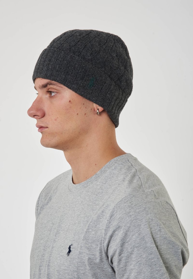 Beanie grigia lavorata a maglia con una texture a coste, con un accento di logo verde. Indossato da una persona con una maglietta grigia chiara, vista di profilo.