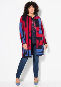 Cardigan ouvert de longueur genou avec des motifs géométriques rouges, bleus et noirs ; associé à un jean foncé et un haut marine. Texture lisse.
