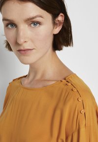 Blusa gialla senape con scollatura rotonda, dettagli con bottoni sulle spalle e tessuto fluido. Design semplice e fascino casual.